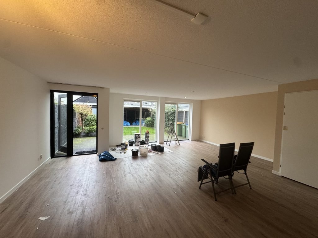 Ruime woonkamer met lichte, gesausde wanden, een houten vloer en tuindeuren, met verfemmers op een loper